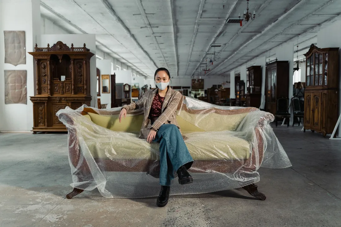 Frau mit Maske sitzt auf einem mit Luftpolsterfolie umhüllten Sofa in einer Möbelhalle