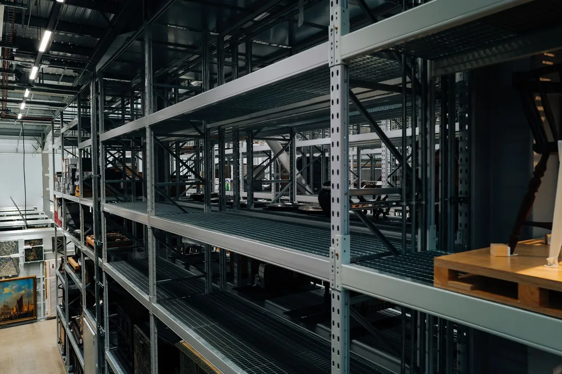 Hohe Metallregale in einer Lagerhalle, darin eingelagerte Bilderrahmen und einzelne Möbelstücke