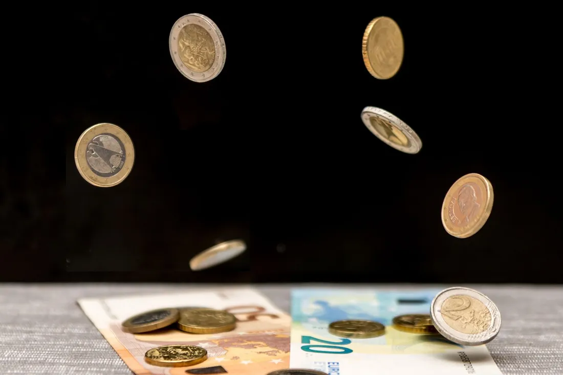 Mehrere Euromünzen in der Luft, darunter Zwanzig-Euro-Schein und einzelne Münzen auf grauem Tischtuch