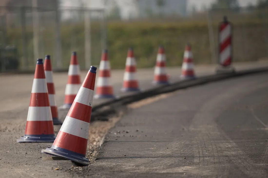 Reihe rot-weißer Leitkegel am Fahrbahnrand, im Hintergrund unscharf eine Leitbake und ein Bauzaun