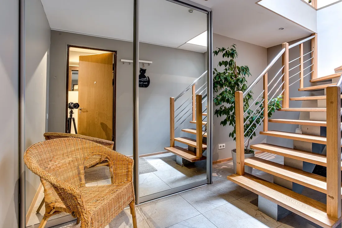 Heller Eingangsbereich mit offener Holztreppe, Spiegelschrank, Korbsessel und Zimmerpflanze auf gefliestem Boden