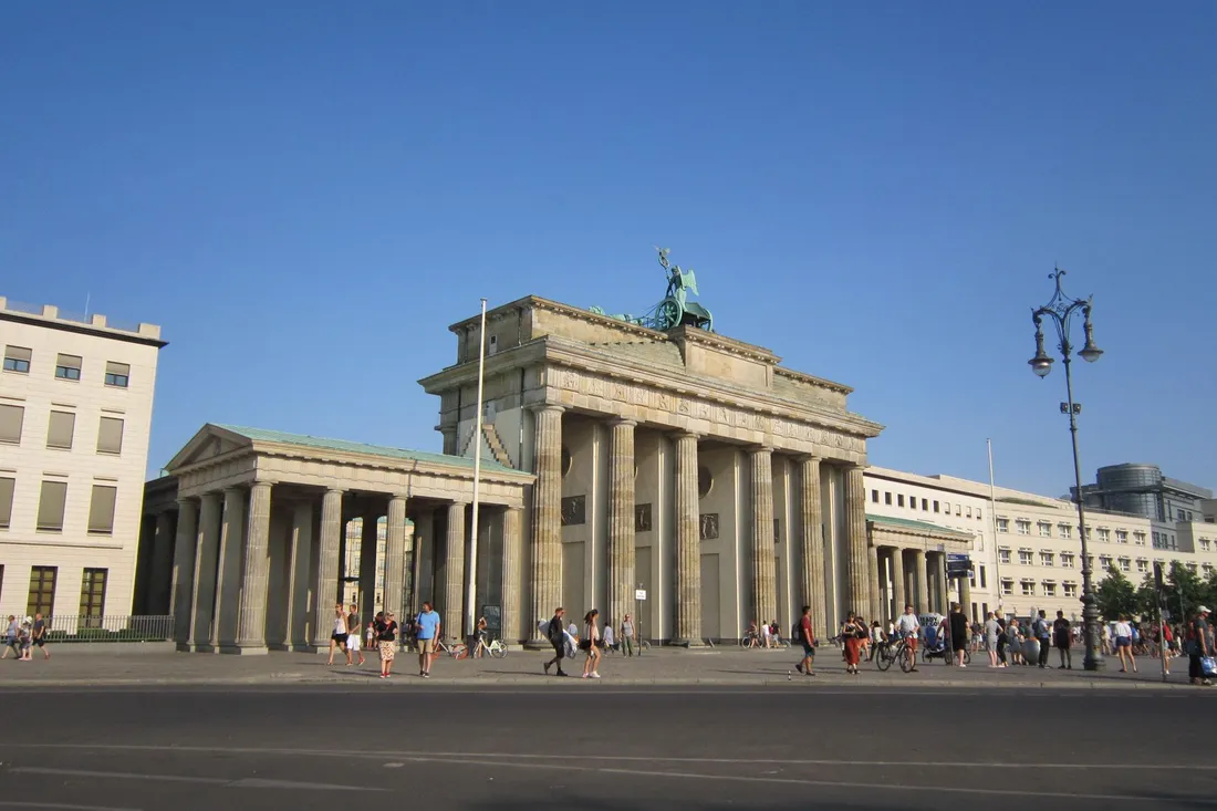 Brandenburger Tor mit Quadriga vor blauem Himmel, davor Passanten auf dem breiten Platz