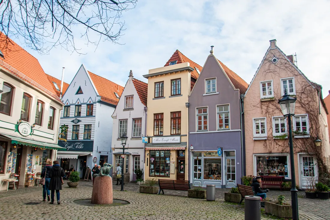 Kleiner Kopfsteinplatz mit bunten Giebelhäusern, Café und einer Bronzefigur auf Sockel