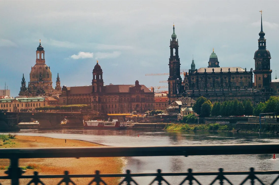 Barocke Kuppeln und Türme hinter einem Fluss, vorn ein schmiedeeisernes Brückengeländer
