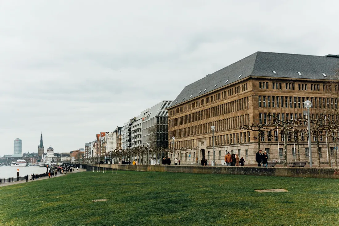 Uferpromenade mit Rasenfläche und großem Sandsteingebäude, dahinter ein spitzer Kirchturm