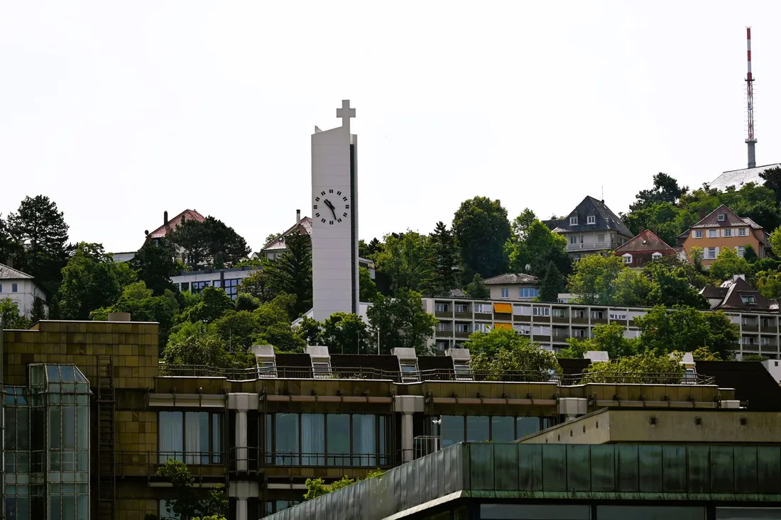 Weißer Kirchturm mit Uhr über bewaldeten Hanghäusern, rechts ein Sendeturm