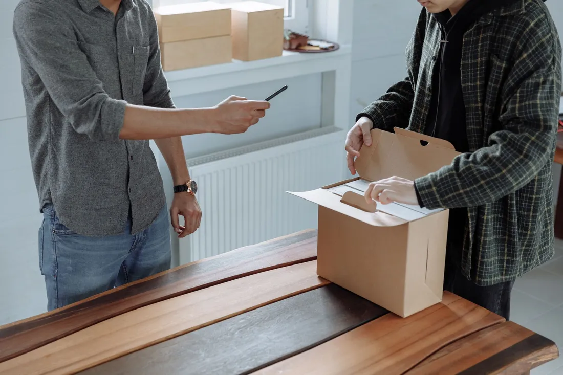 Zwei Männer am Holztisch verschließen einen unbedruckten Karton, einer hält einen Stift