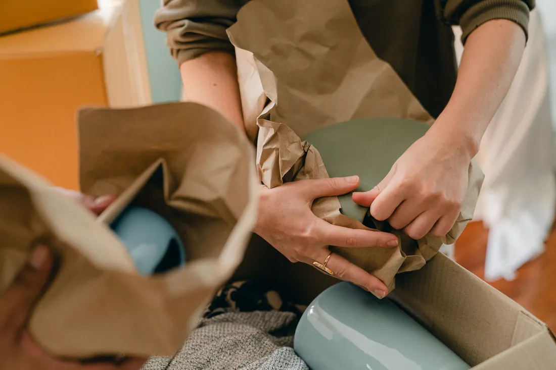 Hände wickeln eine hellblaue Keramikvase in braunes Packpapier über einem offenen Karton